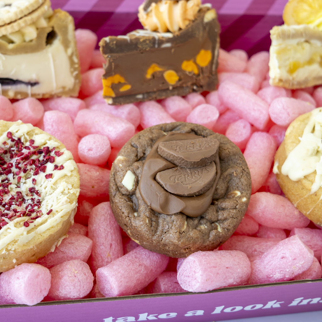 A close-up of assorted baked sweets and chocolates arranged in a pink box. Visible items include a chocolate chip cookie with milk chocolate pieces on top, a slice of chocolate cookie with orange pieces, and other pastries around it.