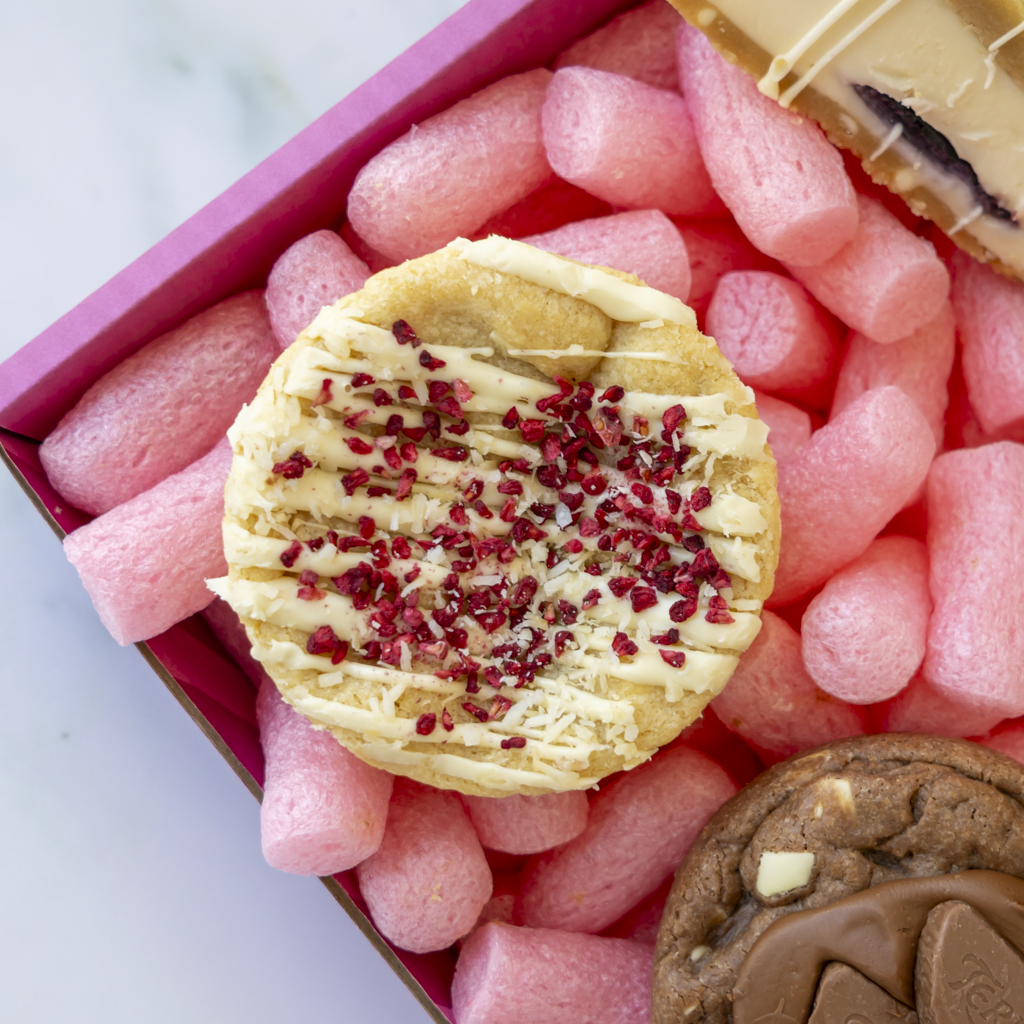 A close-up overhead view of assorted cookies arranged in pink packing foam inside a pink box. The central cookie is round, golden, and topped with white chocolate drizzle and red berry sprinkles.