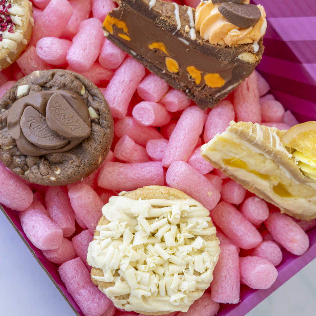 A close-up of assorted gourmet chocolates and cookies arranged in pink packing foam inside a pink box. Visible pieces include a chocolate cake slice with orange drizzle, a chocolate cookie with caramel and chocolate swirls, a white chocolate cookie topped with white chocolate shavings, and a cream-filled pastry with yellow custard.