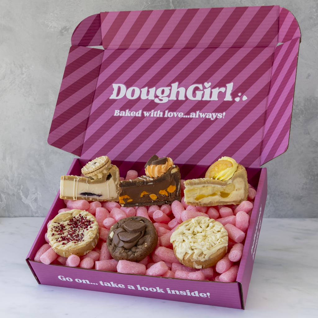 An open pink DoughGirl gift box filled with assorted fruity mini pastries and cakes, set on a light marble surface against a gray wall background.