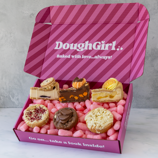 An open pink DoughGirl gift box filled with assorted fruity mini pastries and cakes, set on a light marble surface against a gray wall background.