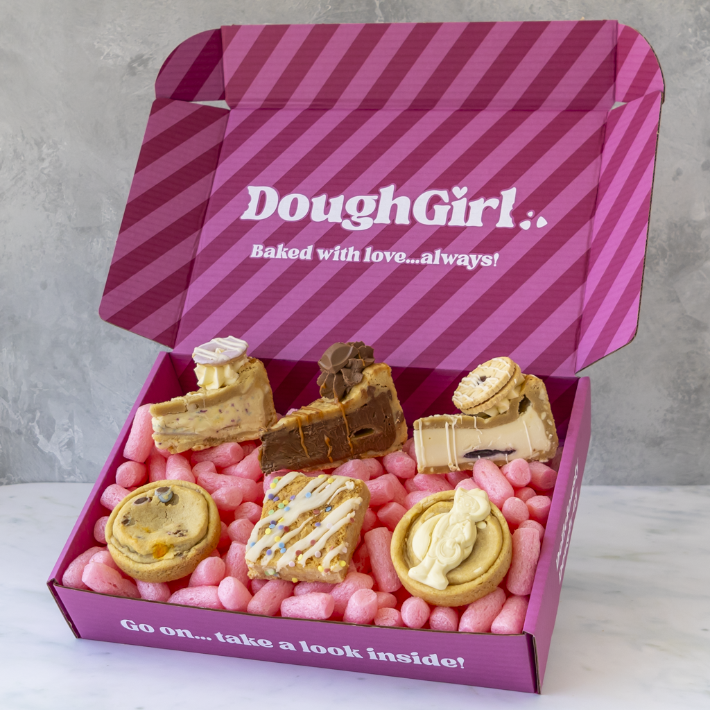 A pink striped DoughGirl baking gift box filled with assorted baked goods, including cakes, pastries, and cookies, set against a light gray marble surface and textured gray background.