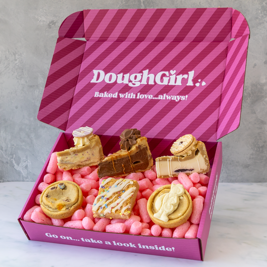 A pink striped DoughGirl baking gift box filled with assorted baked goods, including cakes, pastries, and cookies, set against a light gray marble surface and textured gray background.