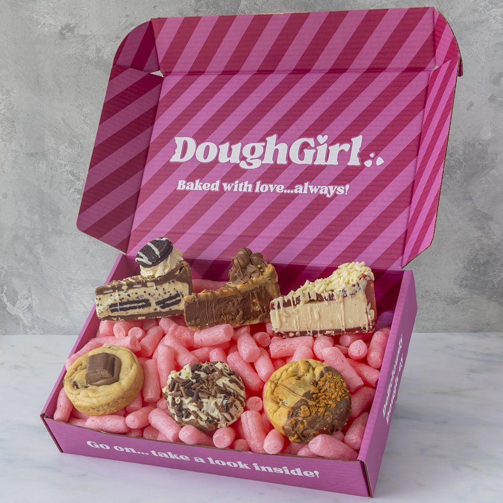 An open pink DoughGirl gift box filled with assorted pastries and cookies on a light marble surface against a gray wall. The box lid has pink diagonal stripes and the brand name 'DoughGirl' printed in white.