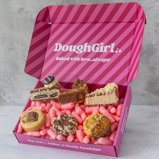 An open pink DoughGirl gift box filled with assorted pastries and cookies on a light marble surface against a gray wall. The box lid has pink diagonal stripes and the brand name 'DoughGirl' printed in white.