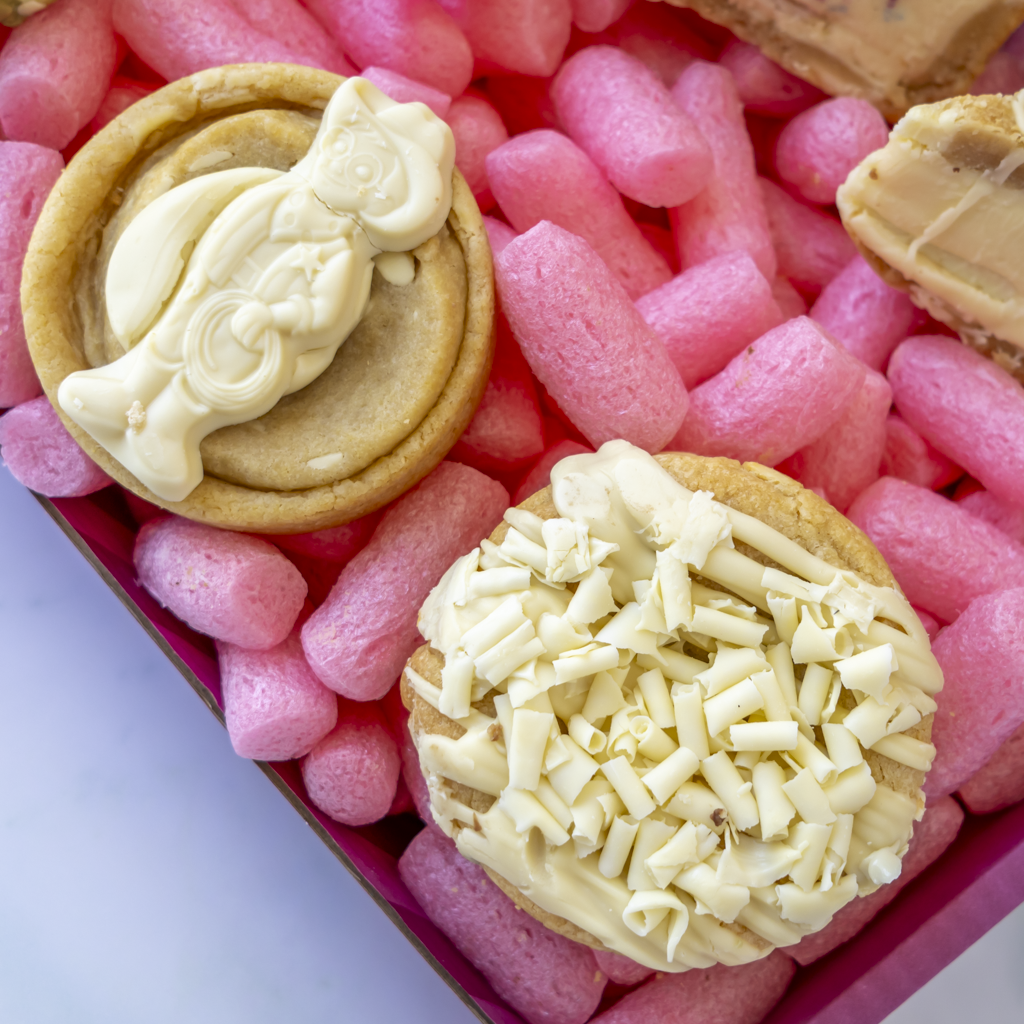 Two decorated cookies in a pink box. One cookie is topped with smooth white chocolate bar Milkybar, and the other is topped with shredded white chocolate.