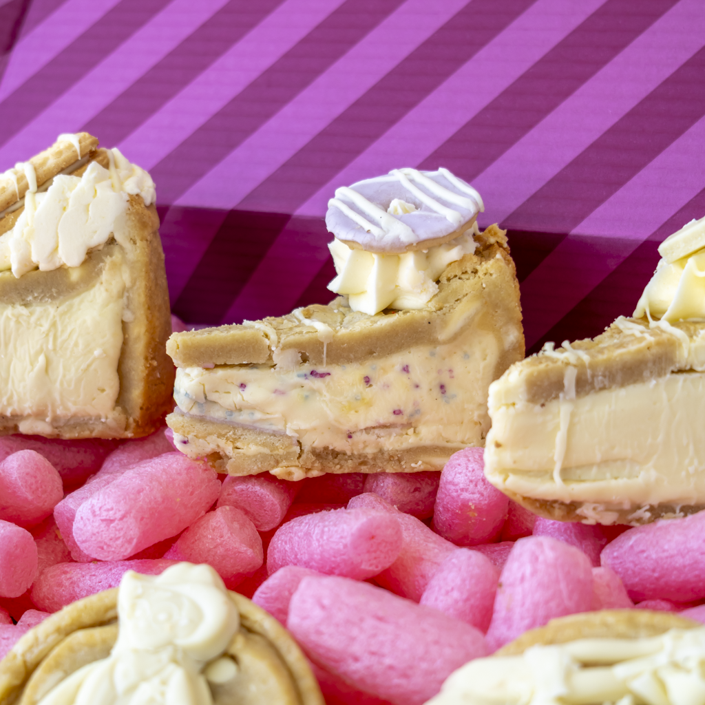 Close-up of several slices of creamy cookie pie with white chocolate drizzle and toppings, arranged on pink candy-coated chocolates against a pink striped background.