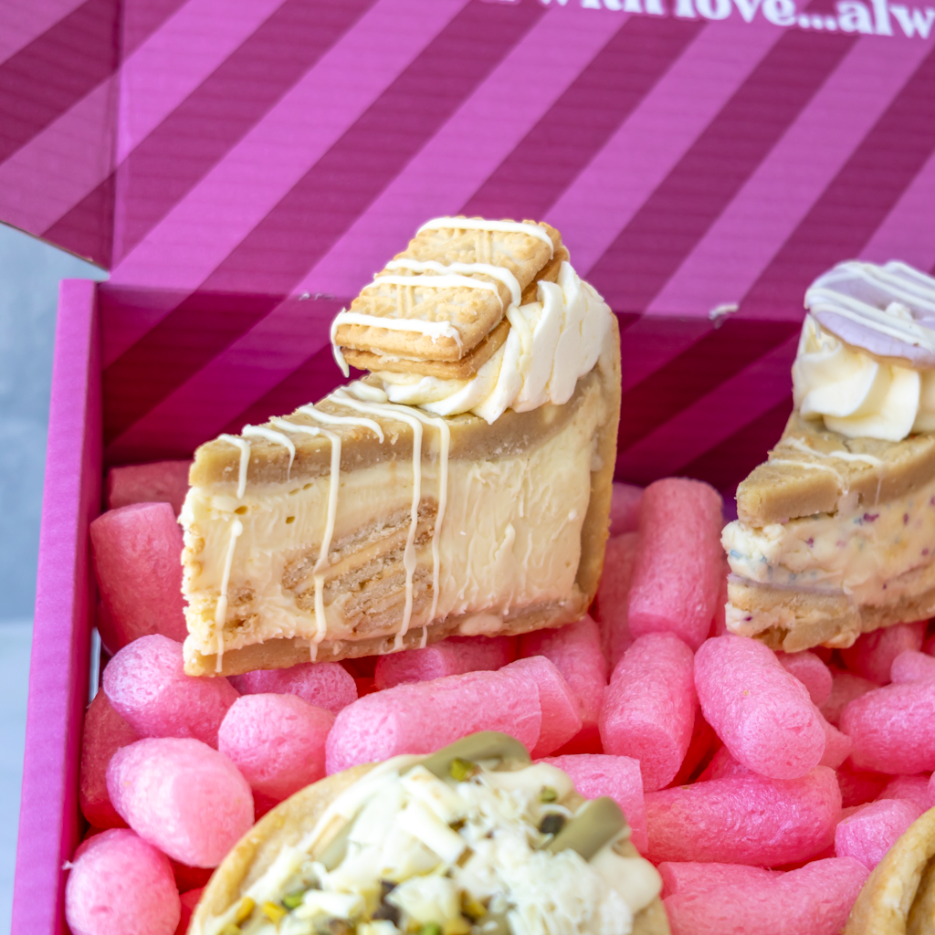 A close-up of assorted gourmet dessert slices displayed inside a pink gift box. The desserts include cookie pie slices with white drizzle and cookie toppings, and other sweet pastries and cookies.