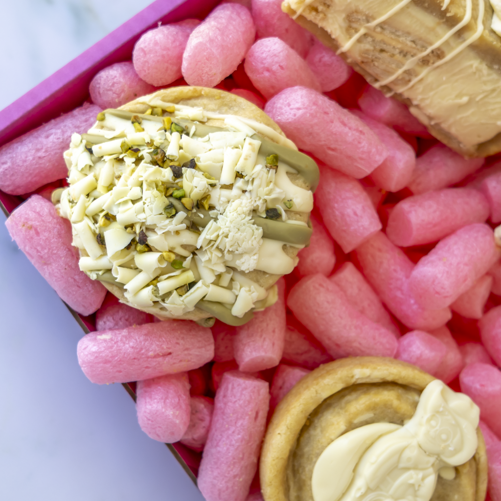 A close-up overhead view of a pink gift box featuring two decorated cookies and part of a wrapped item. The main visible product is topped with pistachio and white chocolate.