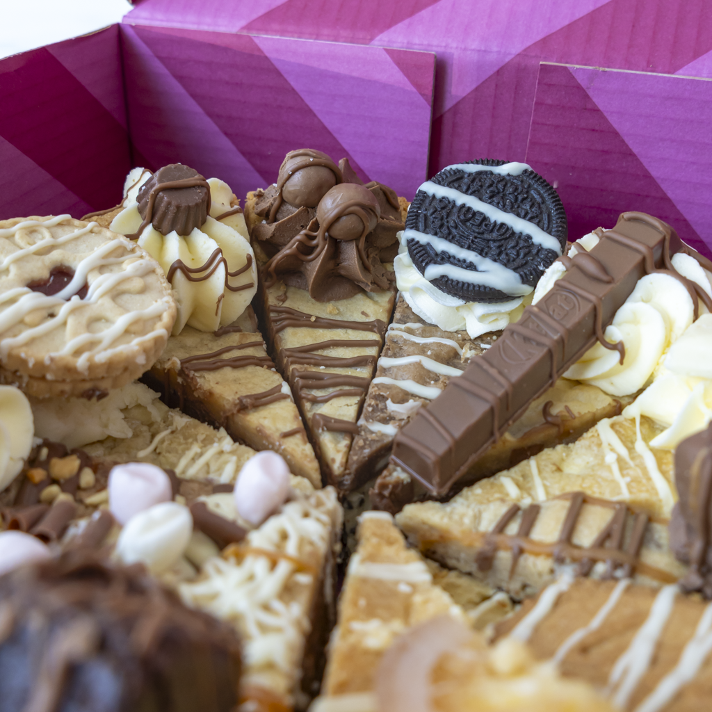 A close-up of assorted chocolate and cookie bars arranged in a box with a pink geometric patterned lid. The bars include chocolate drizzle, cream frosting, Oreo cookies, and chocolate-covered treats.
