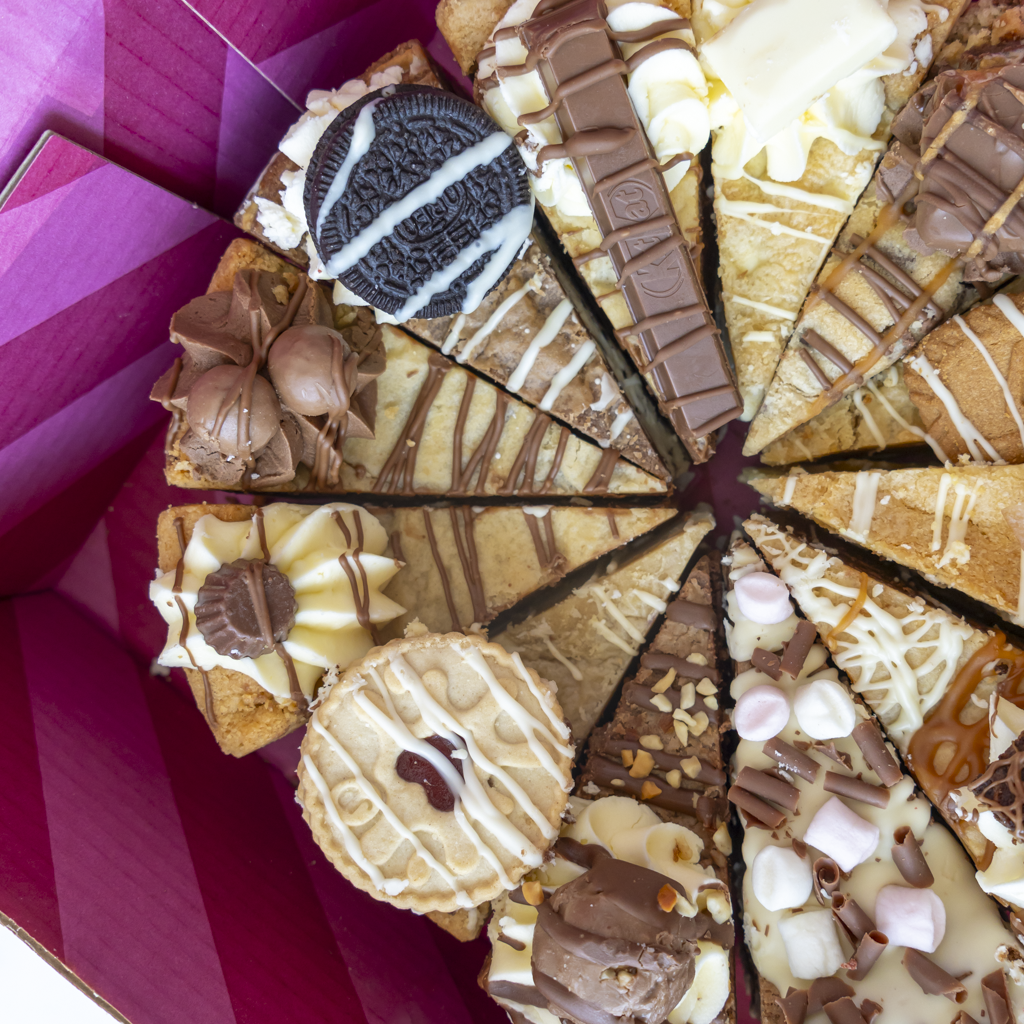 A close-up overhead view of a round chocolate cake or dessert pie sliced into wedges, topped with white chocolate drizzle, chocolate chunks, nuts, marshmallows, and a whole Oreo cookie, presented inside a pink box.