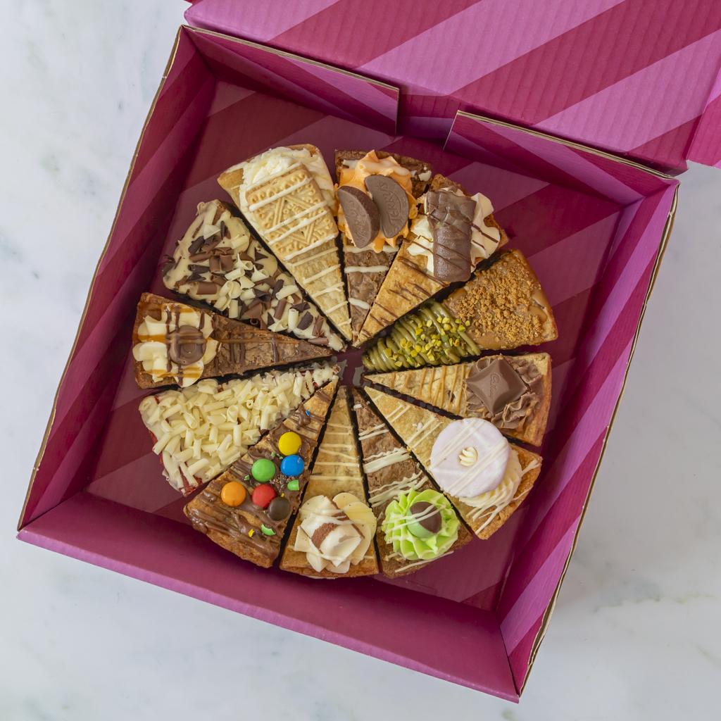 A circular assortment of chocolate and pastry slices presented inside an open pink gift box on a light marble surface.