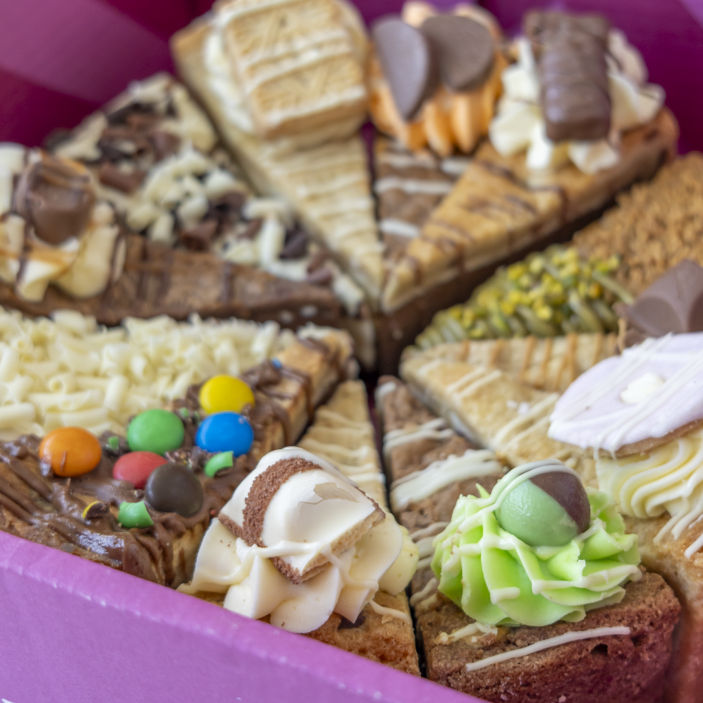 A close-up of assorted chocolate and cream cookie pie slices arranged in a pink container, including cake pieces topped with chocolate, white chocolate drizzle, M&M's candies, green frosting, and chocolate pieces.