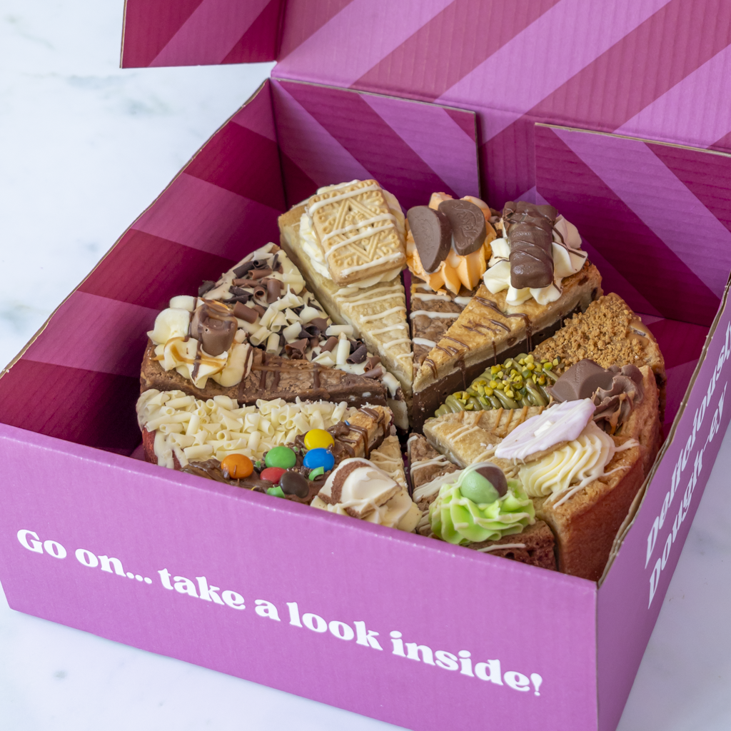 A pink gift box filled with assorted chocolate and cookie pie slices, photographed from above. The box lid is open and printed with the phrase 'Go on... take a look inside!'
