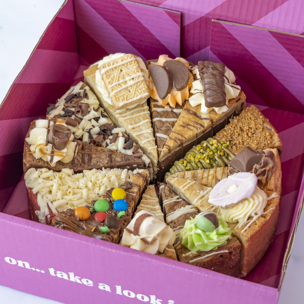 A boxed assortment of dessert slices presented in a pink box. The cake-like dessert pieces are decorated with chocolate, cream, nuts, sprinkles, and green frosting, arranged in circular segments inside a magenta package.