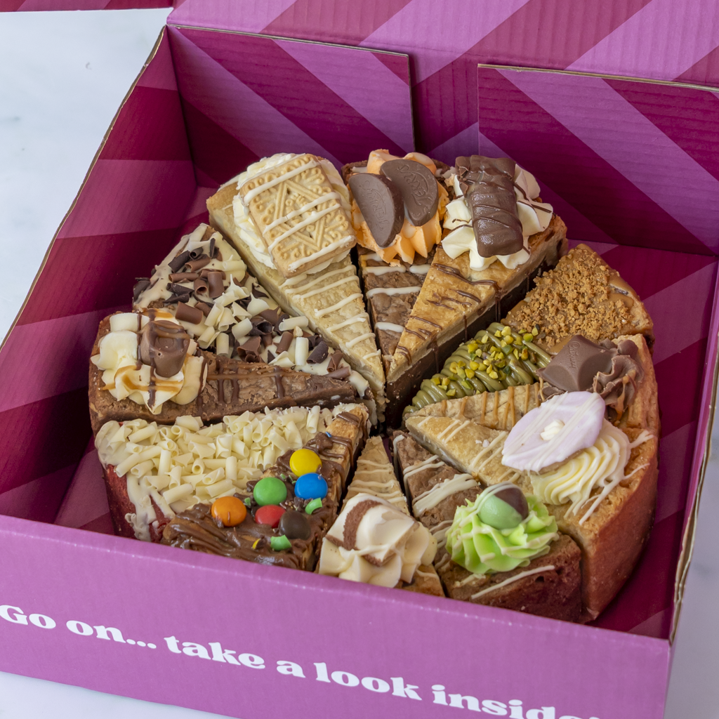 A round cookie pie presented inside a pink gift box, sliced into sections decorated like different flavours of dessert, with chocolate, cream, nuts, sweets, and green frosting details. The box has a pink geometric pattern and the text 'Go on...take a look inside'