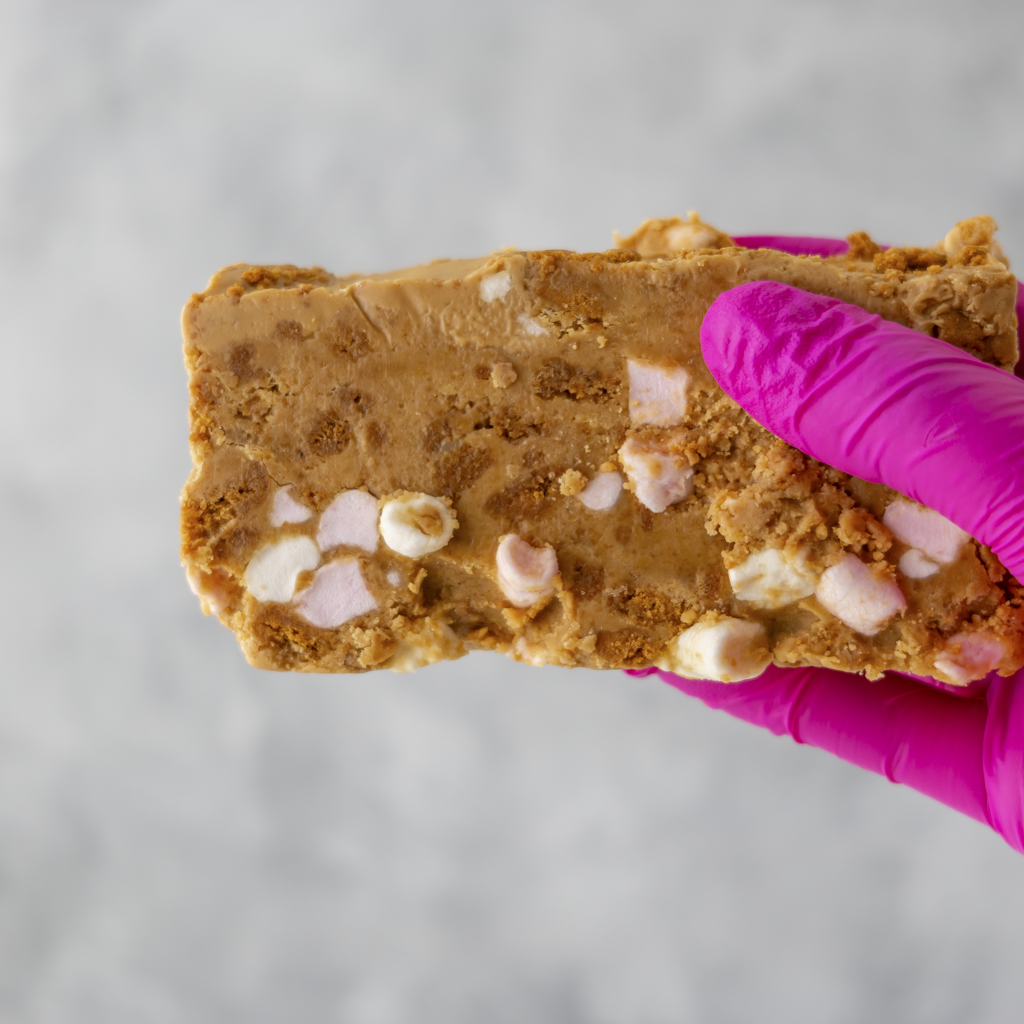 Hand holding a Biscoff Rocky Mountain Road slab with marshmallows and chocolate chips, wearing a pink glove on a gray background