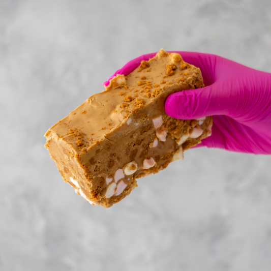 Hand holding a piece of Biscoff Rocky Mountain Road slab with a pink glove on a gray background