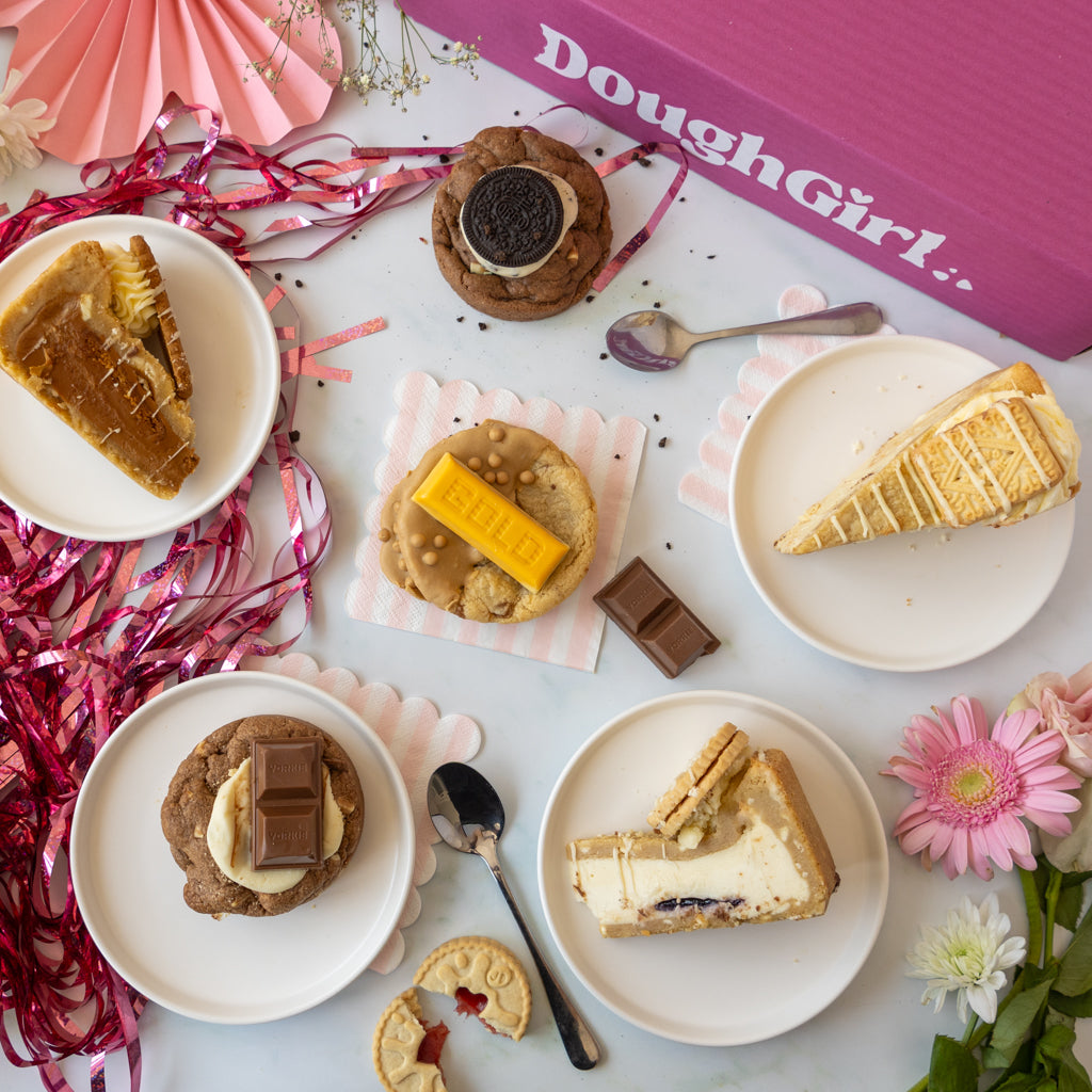 A styled flat-lay of assorted baked goods on white plates, including slices of cookie pie and cookies, arranged with pink paper fans, tinsel, flowers, and a pink DoughGirl box in the background.