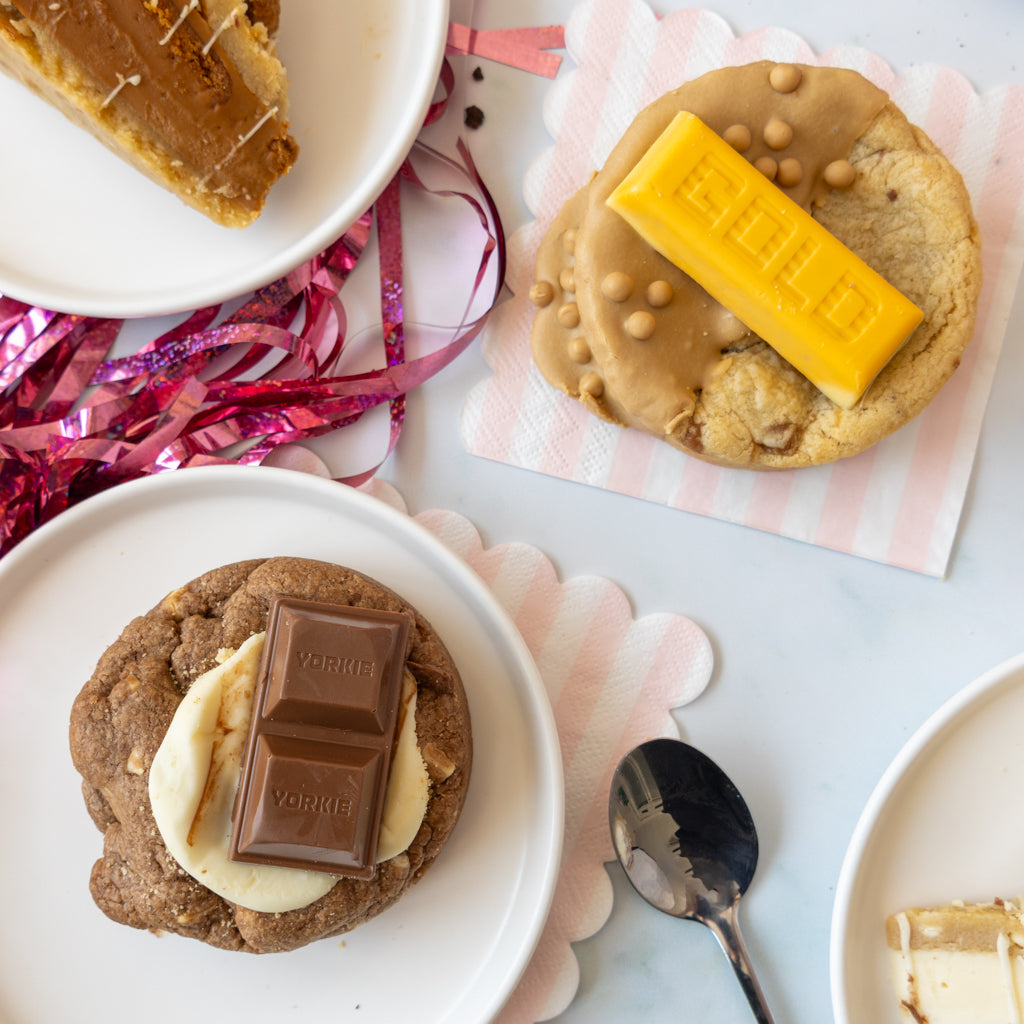 Three chocolate chip cookies decorated with various toppings, including a yellow chocolate bar, a milk chocolate bar, and banana slices, arranged on a white surface with pink and white scalloped paper and a spoon.
