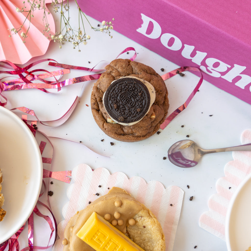A styled flat-lay of two gourmet cookies on a white surface with pink and white paper fans, ribbons, a spoon, and a pink box labeled 'DoughGirl'. The top cookie is chocolate chip with an Oreo cookie on top, and the bottom cookie is with a yellow gold bar on top.