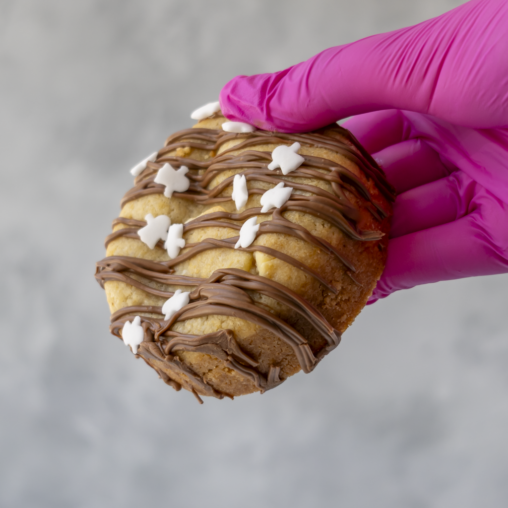 A hand wearing a bright pink glove holds a Boomallow Deluxe Cookie topped with drizzled chocolate and small white ghost sprinkles, against a soft grey background.