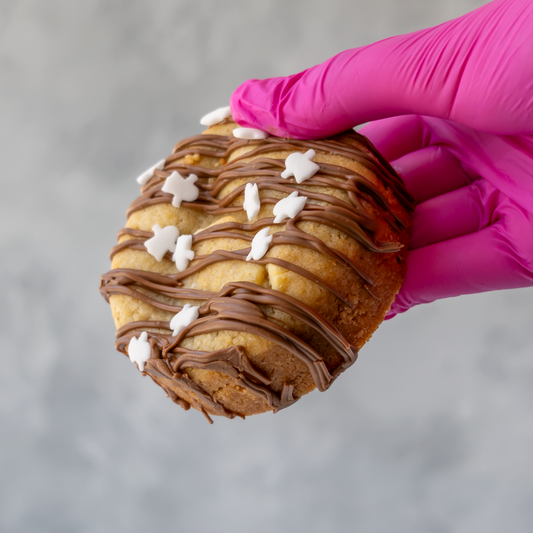 A hand wearing a bright pink glove holds a Boomallow Deluxe Cookie topped with drizzled chocolate and small white ghost sprinkles, against a soft grey background.