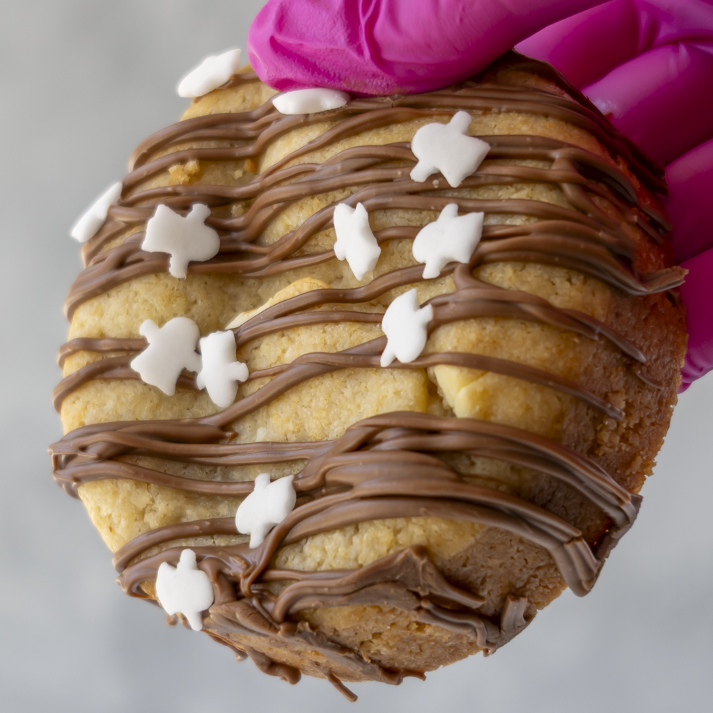 A close-up of a round cookie topped with chocolate drizzle and small white ghost icing shapes, held by a bright pink gloved hand against a light gray background.
