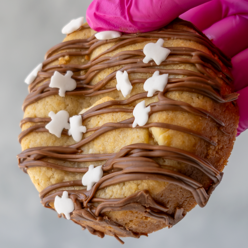 A close-up of a round cookie topped with chocolate drizzle and small white ghost icing shapes, held by a bright pink gloved hand against a light gray background.