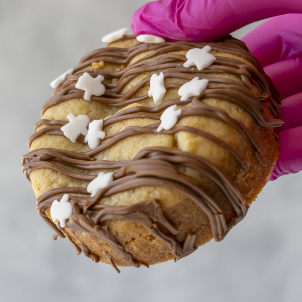 A close-up of a DoughGirl Boomallow Deluxe Cookie topped with chocolate drizzle and small white ghost decorative pieces, held by a hand wearing a bright pink glove against a soft gray background.