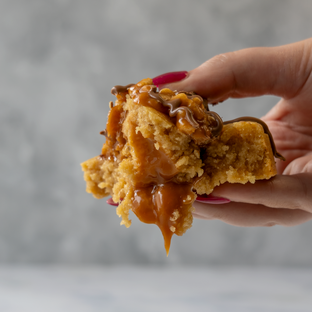 Hand holding a piece of Lion Bar Deluxe Cookie with caramel sauce against a gray background