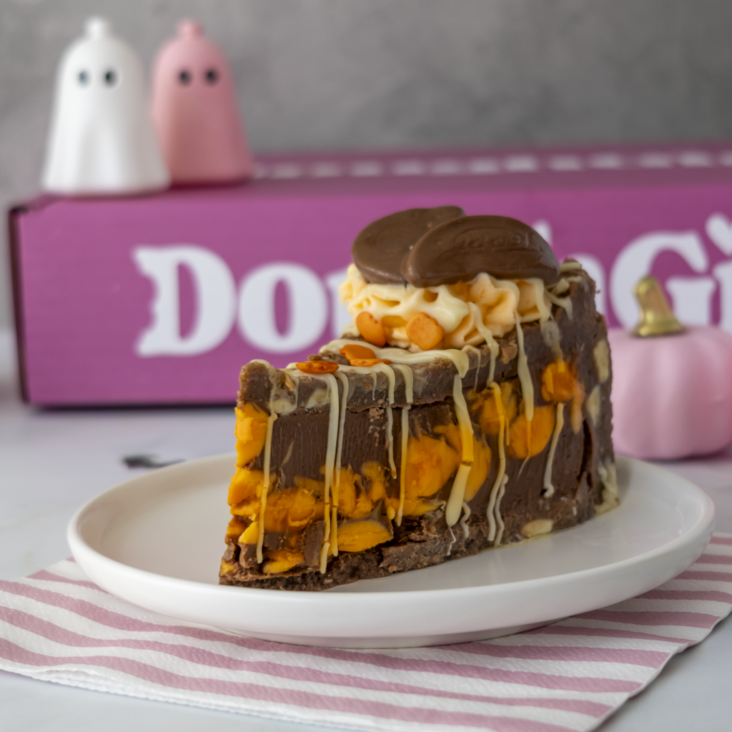 A slice of chocolate and orange treat pie on a white plate, decorated with white drizzle and topped with a chocolate wafer cookie. The background features a pink box with white text and small ghost figurines.