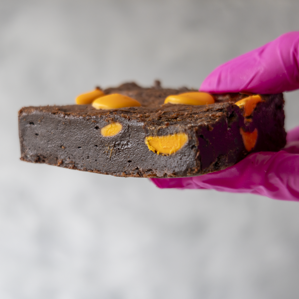 Chocolate brownie with orange sweets held by a pink gloved hand against a light gray background
