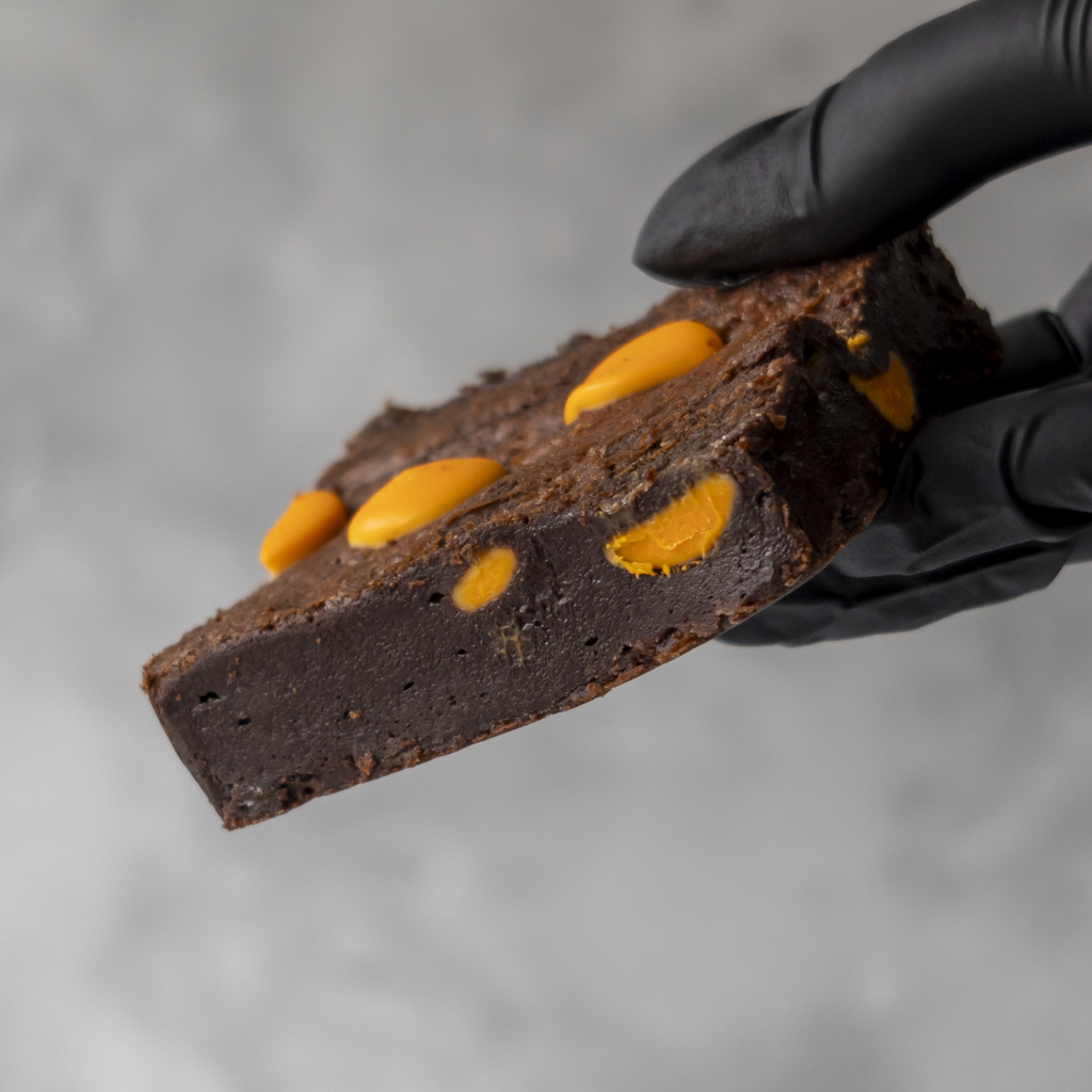 Chocolate Orange Brownie Slab with orange chips held by a black gloved hand against a neutral background