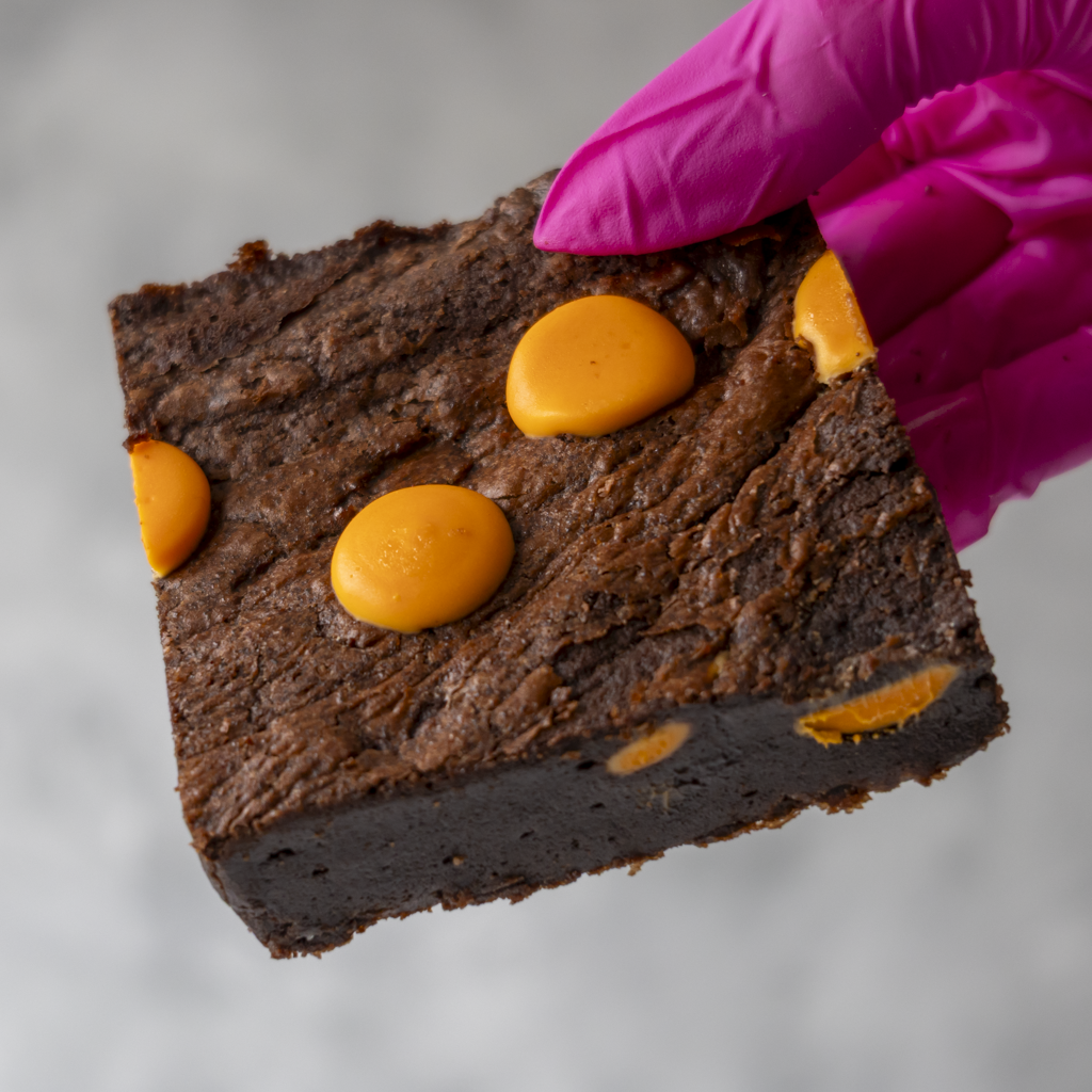 Chocolate Orange Brownie Slab by DoughGirl with orange sweets held by a gloved hand on a neutral background