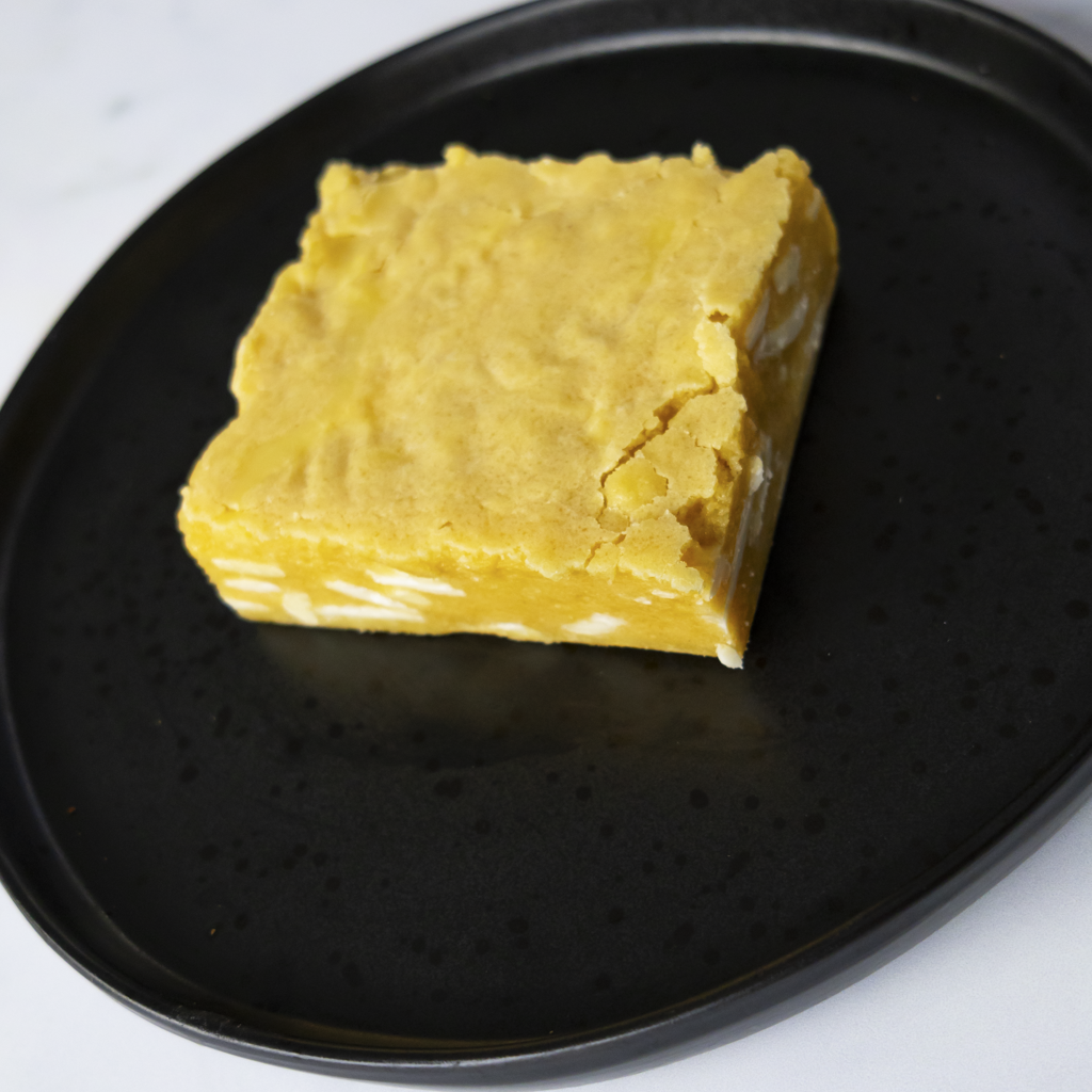 Square Classic Blondie Slab, made with real vanilla, golden brown sugar, and creamy white chocolate chunks, on a black plate