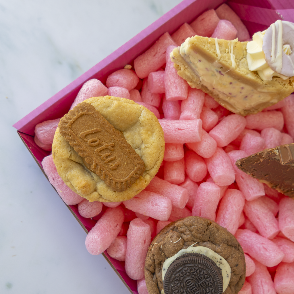 A close-up of assorted sweets arranged in a pink box filled with pink packing foam. Visible items include a round biscuit stamped with 'Lotus', a slice of cheesecake with drizzle and a white chocolate swirl, a chocolate cookie with an Oreo cream filling, and another chocolate cookie piece.