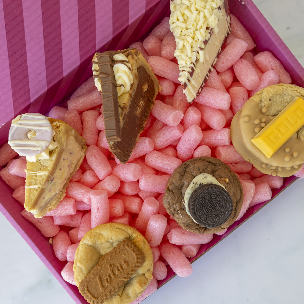 A pink gift box filled with pink packaging, containing assorted dessert-shaped items including chocolate cake slices, cookies, and a biscuit with cream and chocolate.