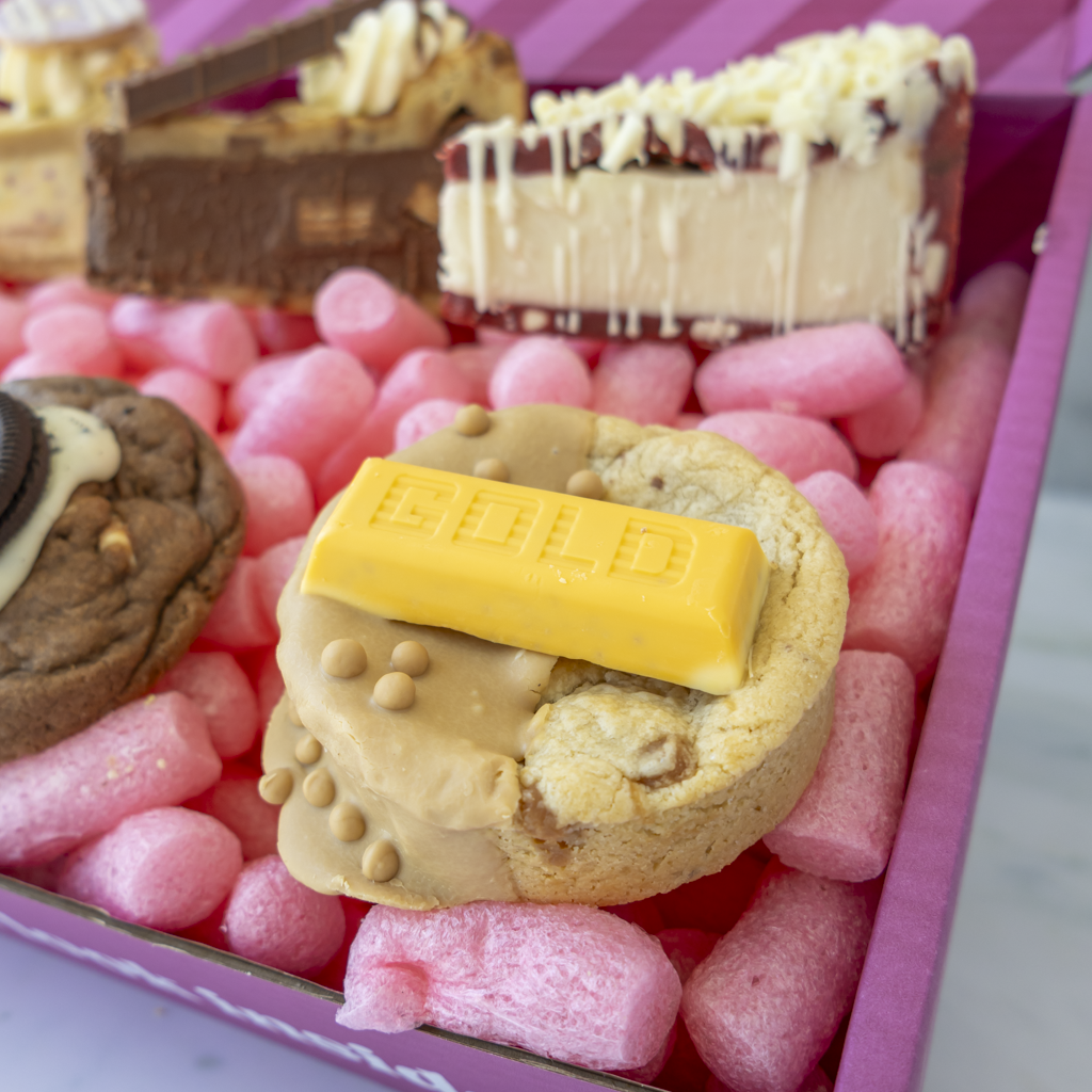 A close-up of a boxed dessert gift set filled with pink packing foam. The set includes a chocolate chip cookie topped with a yellow gold candy bar, a red velvet chocolate cake slice with white drizzle, a brown chocolate cake slice, and a cookie with cream and Oreo-style topping, all arranged in a pink box.