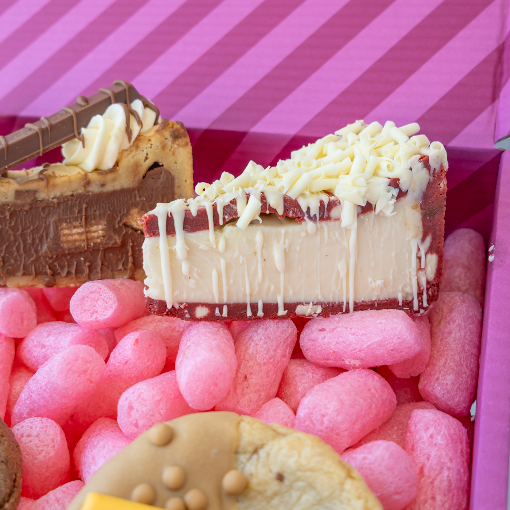 A close-up of assorted baked goods arranged in a pink box. Visible items include a slice of red velvet cookie pie topped with white chocolate drizzle, a chocolate cake slice with whipped cream, a cookie, and more pink candy-coated pieces.
