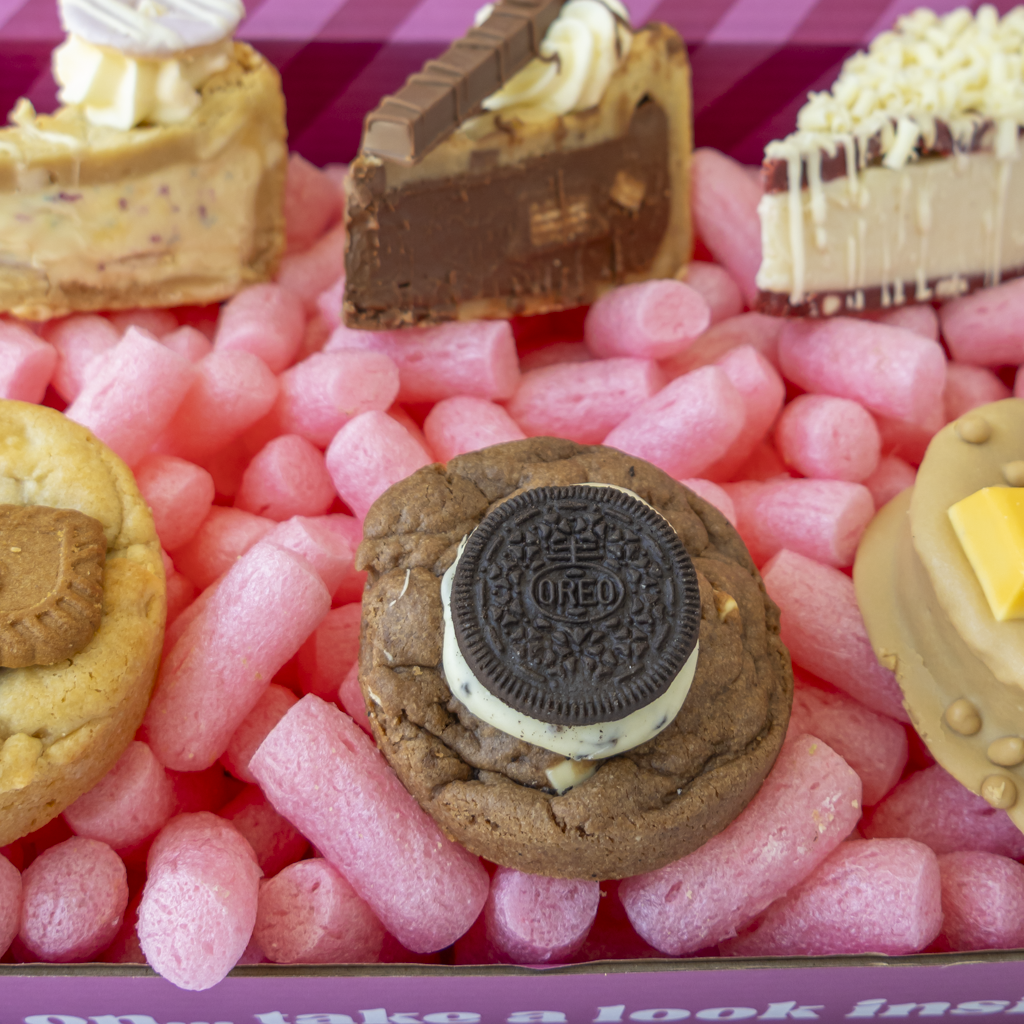 A close-up of assorted gourmet sweets arranged in pink packing peanuts inside a box. Visible items include a chocolate cookie with an Oreo cookie on top, a milk-chocolate slice with cream swirl, a cream-drizzled dessert, and other baked goods.