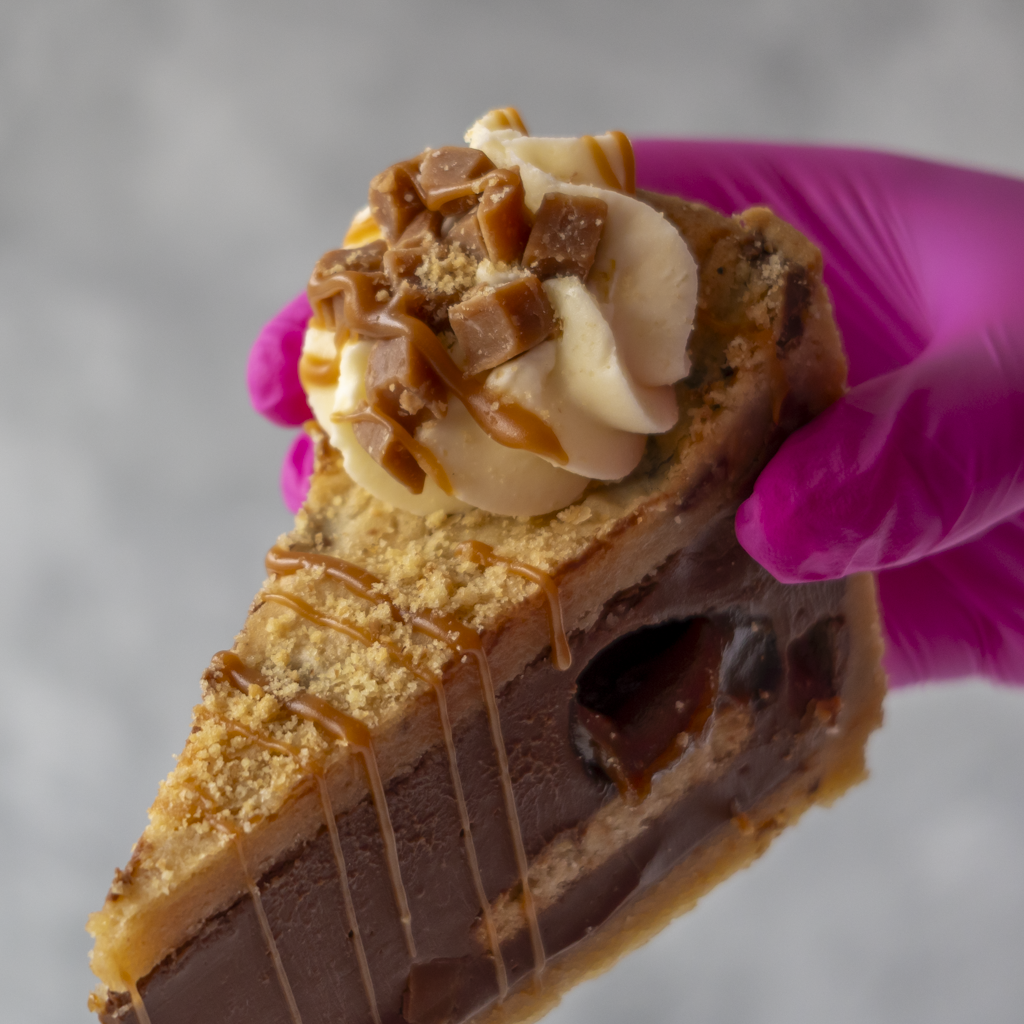 A close-up of a hand wearing a bright pink glove holding a slice of chocolate cookie pie with whipped cream, caramel drizzle, and chocolate chunks on top, against a soft gray background.