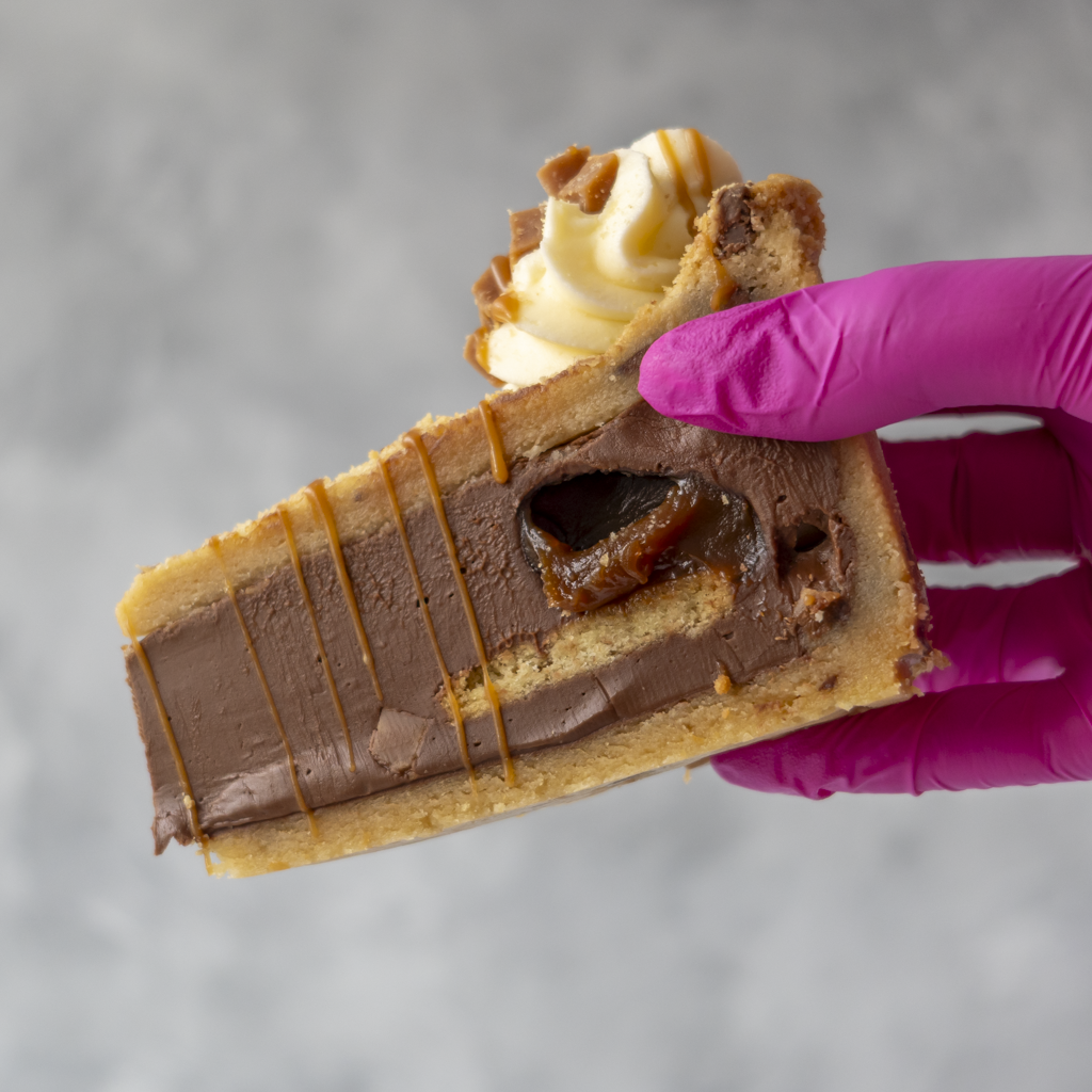 A close-up of a hand wearing a bright magenta glove holding a slice of layered chocolate and caramel cookie pie slice with whipped cream on top, set against a soft gray background.