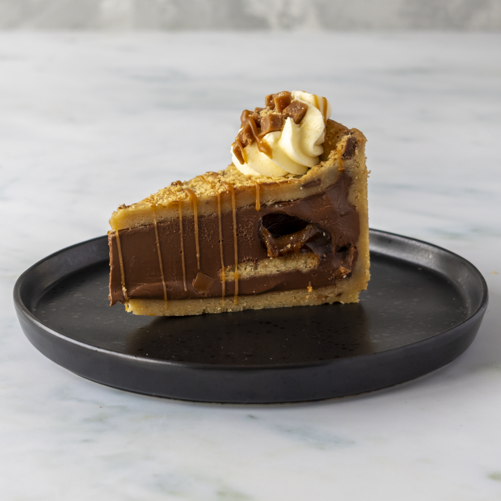 A slice of gourmet Banoffee Cookie Pie on a round black plate, featuring a chocolate and caramel filling with a cracker crust and whipped cream on top, set on a light marble surface.