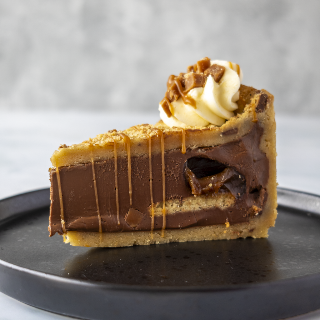A slice of chocolate pie with caramel drizzle and a whipped cream topping, served on a dark plate against a light gray background.