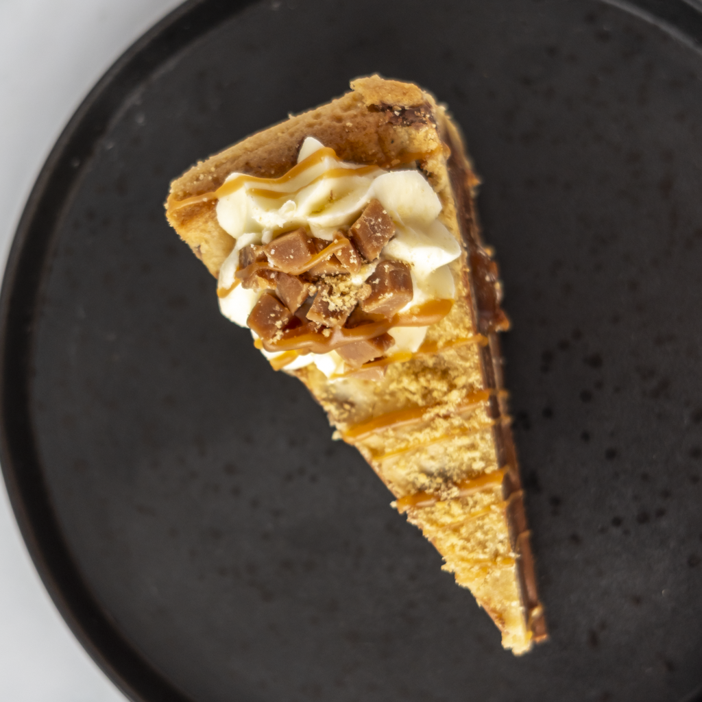 A single slice of gourmet cookie pie on a dark speckled plate, topped with whipped cream, caramel drizzle, and chocolate cookie crumbles.