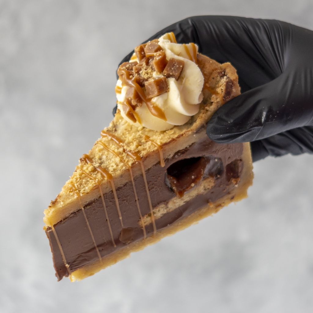 A slice of rich chocolate pie with caramel drizzle, whipped cream, and caramel topping, held by a hand wearing a black glove against a light gray background.