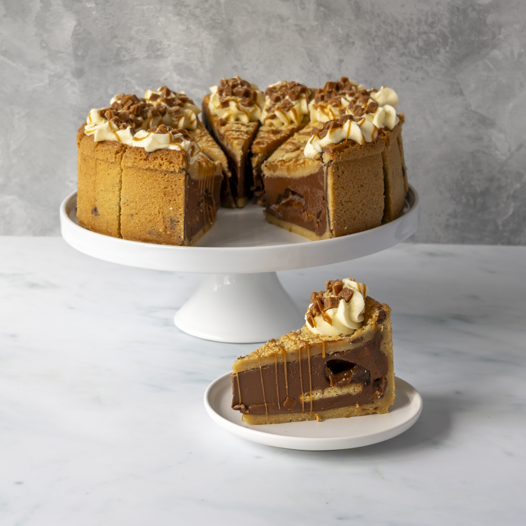 A layered Banoffee Cookie Pie Slice with chocolate and caramel filling, served on a white cake stand with a single slice on a small plate in front, against a gray textured background.