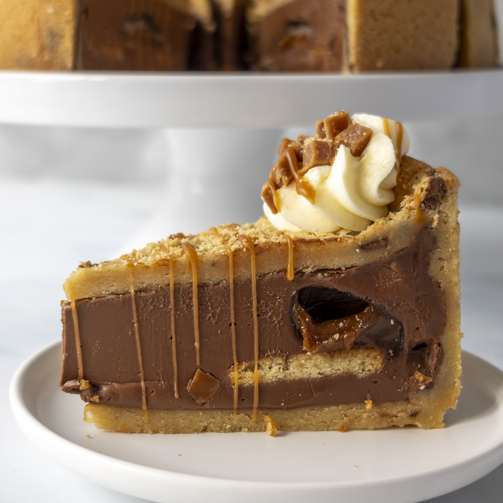 A slice of chocolate cookie pie on a white plate, topped with whipped cream and caramel drizzle, with a blurred background showing more pie slices on a serving stand.