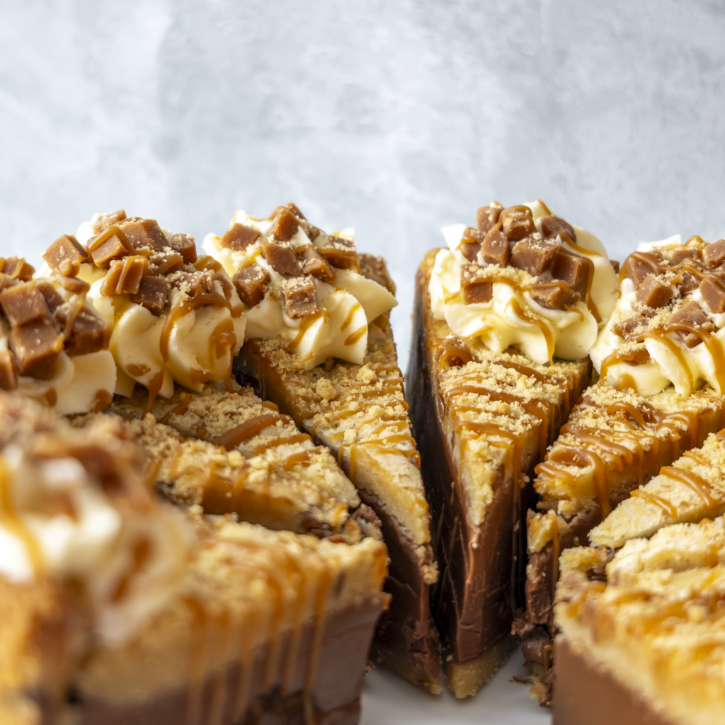 Close-up of several slices of a DoughGirl Banoffee Cookie Pie Slice, showing creamy vanilla or caramel cheesecake layers, chocolate chunks, whipped cream, and topping with caramel drizzle and cookie crumbs, arranged on a white plate against a light gray background.